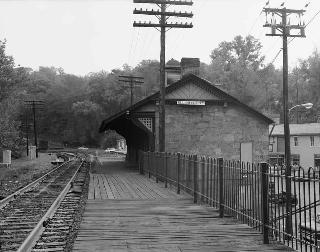 This black and white photo shows the Ellicott Station next to the railroad tracks with buildings and trees in the background.