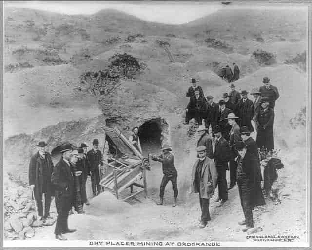 Black and white photograph of two miners at the entrance to a tunnel, surrounded by a group of visiting men in suits.