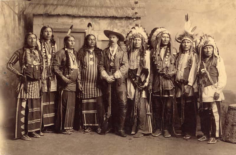 This sepia photo shows Buffalo Bill Cody and a number of Native Americans all in regalia