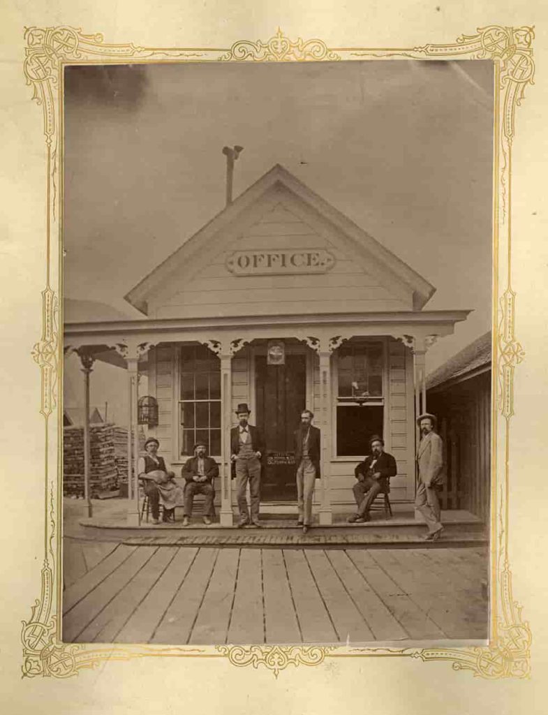 Sepia-toned photograph of a small Victorian-style office building with six men on the porch.